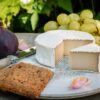 Camembert style vegan cheese by Mouse's Favourite photographed on a late summer's day showing a small segment in front and the rest of the cheese wheel behind with a fig and fruity, chunky crackers on the side